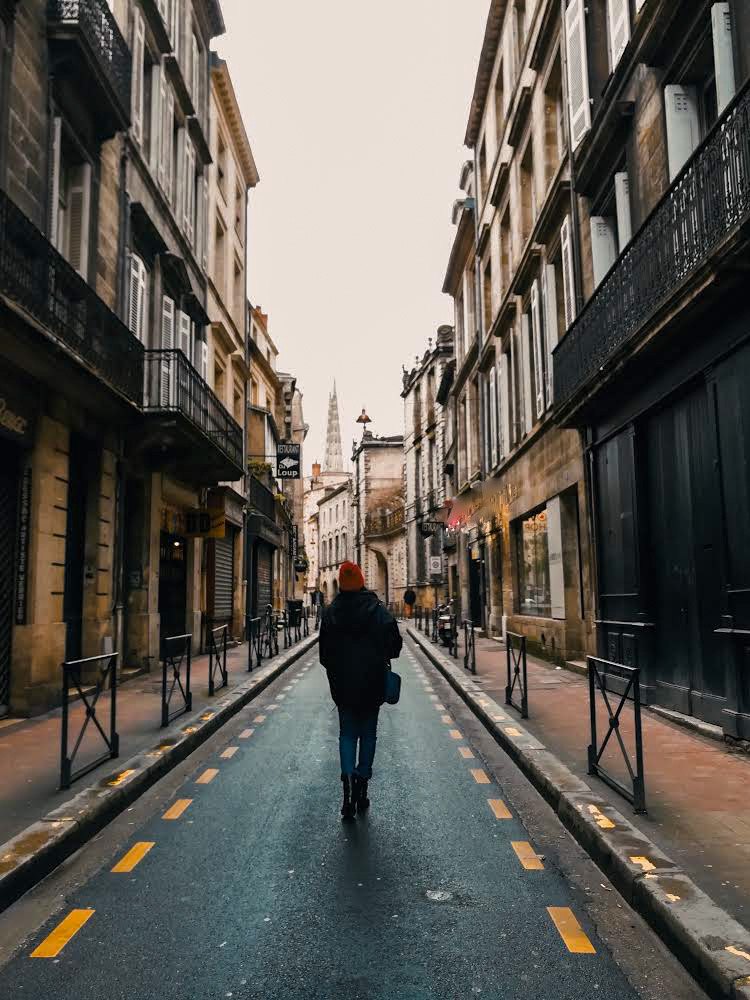 A narrow European street lined with stone buildings and balconies, Ayesha walking away down the middle