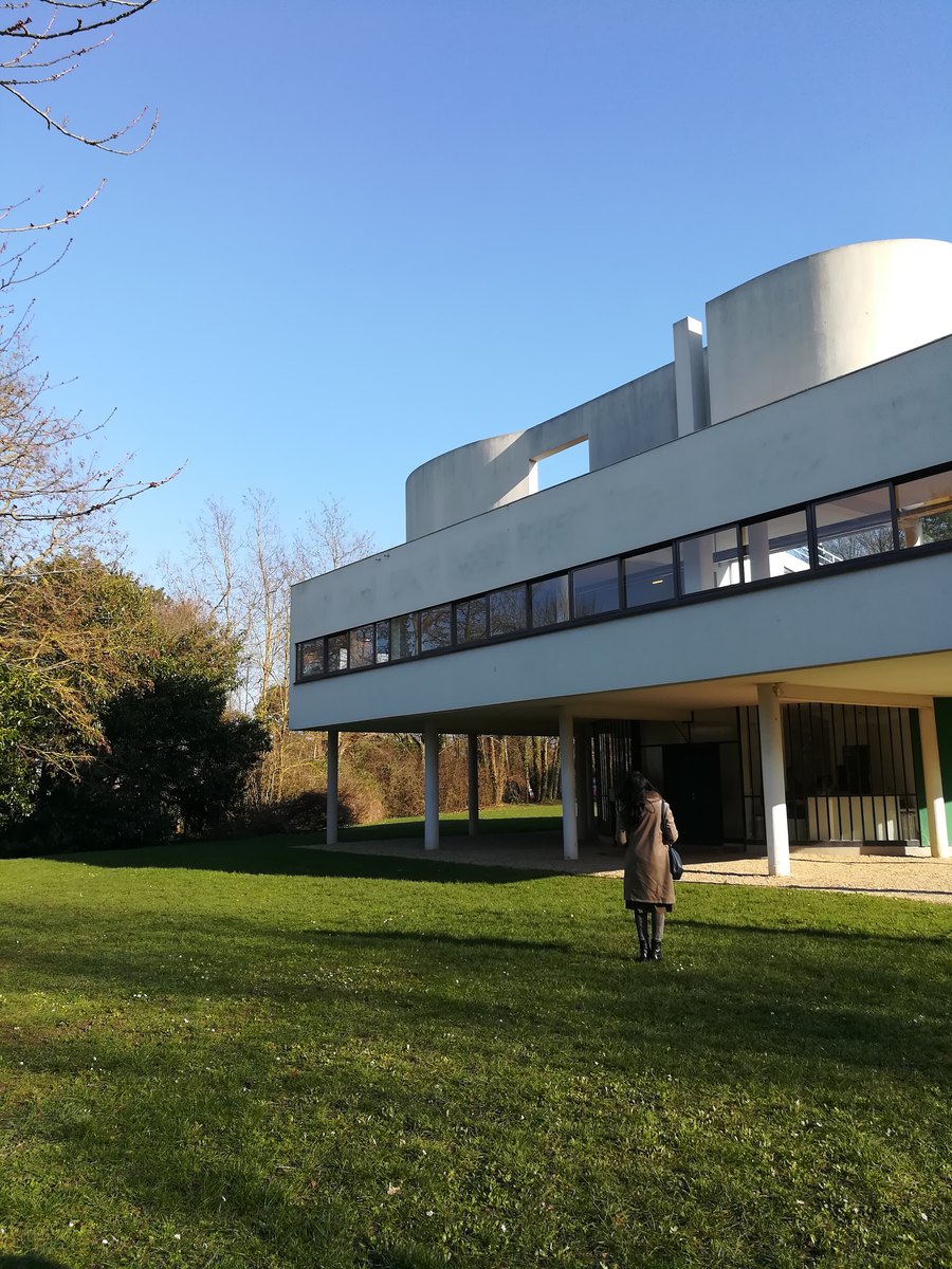 A white modernist villa raised on slender columns above a sunny lawn, Ayesha looking up at it